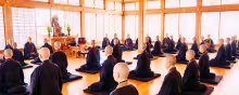A group of nuns and monks in robes sitting Zazen in a brightly lit meditation hall. 
